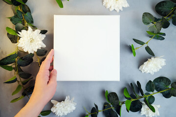 Female hand holding blank canvas frame, white flowers and plant branches on background. Wrapped white canvas for mockup poster or romantic message. Spring, summer concept. Copy space. Top view.