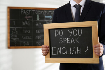 Teacher holding small chalkboard with inscription Do You Speak English? in classroom, closeup