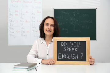 Teacher holding small chalkboard with inscription Do You Speak English? at table in classroom