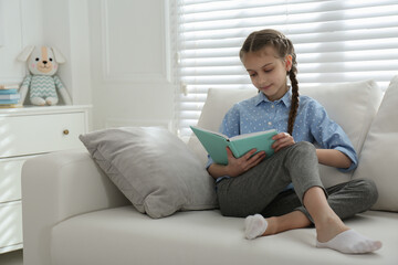 Fototapeta premium Cute little girl reading book on sofa at home