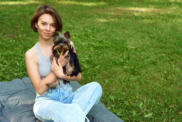Naklejka na ściany i meble Cute beautiful woman with little Yorkshire Terrier in a park outdoors. Lifestyle portrait