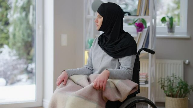Portrait of frustrated desperate Muslim woman sitting in wheelchair in living room sighing. Sad Middle Eastern young handicapped lady at home indoors. Disability and paralysis