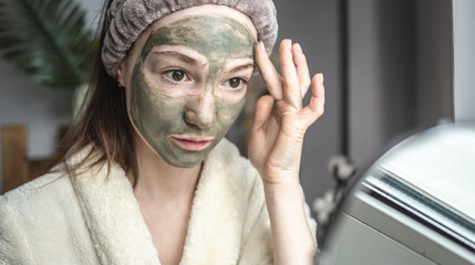 Young beautiful woman in a dressing bathrobe is applying a green cosmetic mask to her face. Concept of skin care and lifestyle