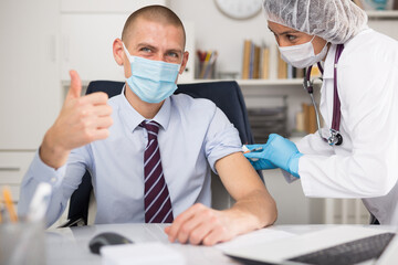 Doctor in white coat and protective mask giving vaccine injection to manager in office