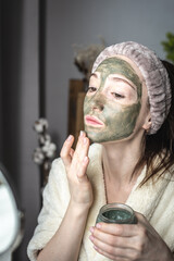 Young pretty woman in a bathrobe in front of a mirror is applying a green cosmetic mask to her face. Concept of skin care, the use of natural cosmetics and lifestyle