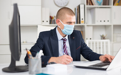 Young man entrepreneur in protective face mask working alone on laptop at his workplace