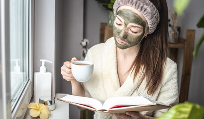 Pretty young woman in a bathrobe and with a green cosmetic mask on her face is reading a book and...