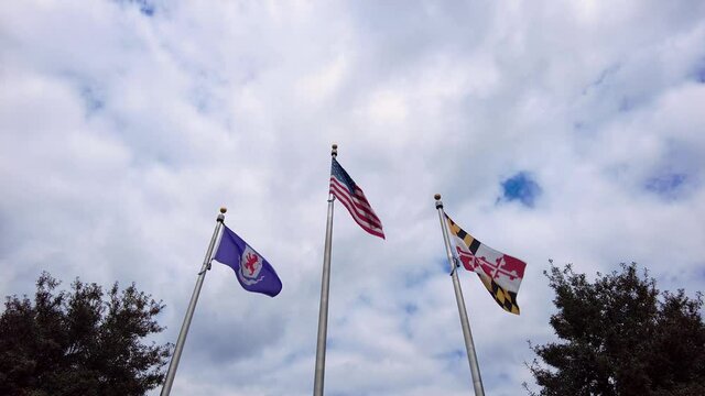Flags of the USA, State of Maryland, and Talbot County, MD are waving on side by side flag posts on a cloudy day.