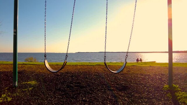 Two empty swings at a waterfront playground are moving gently in the wind at sunset with reflection of sunlight in Chesapeake Beach seen in Background. Versatile footage taken at eastern shore, md