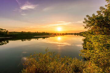 Obraz na płótnie Canvas Scenic view at beautiful summer river sunset with reflection on water with green bushes, grass, golden sun rays, calm water ,deep blue cloudy sky and glow on a background, spring evening landscape
