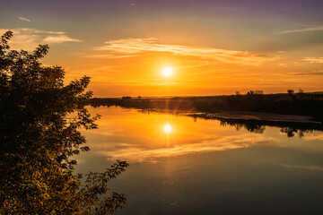 Scenic view at beautiful summer river sunset with reflection on water with green bushes, grass, golden sun rays, calm water ,deep blue cloudy sky and glow on a background, spring evening landscape