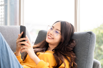 Happy young asian woman relaxing at home she is sitting on sofa and using mobile smartphone