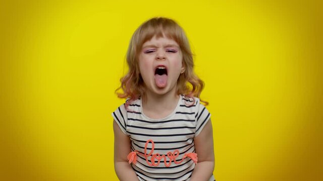 Portrait of cheerful funny blonde child girl 5-6 years old showing tongue making faces at camera, fooling around, joking, aping with silly face, teasing. Yellow studio background. Teen kid children