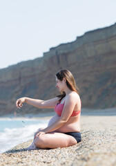 Fototapeta na wymiar Pregnant woman in bikini resting on pebble beach in early time