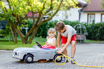 Two happy children playing with big old toy car in summer garden, outdoors. Kid boy refuel car with little toddler girl, cute sister inside. Boy using garden hose and fill up with gasoline, sibling