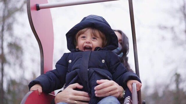 Baby toddler having fun at playground with mother during covid-19 winter season. Child sliding down tobogan