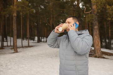A young man in a gray jacket drinks coffee and talks on the phone in a snowy forest.