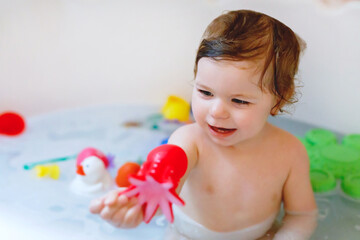 Cute adorable baby girl taking foamy bath in bathtub. Toddler playing with bath rubber toys.