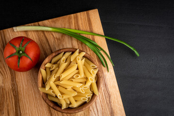 Raw pasta in wooden bowl with cheese, tomato and green onion on black background.