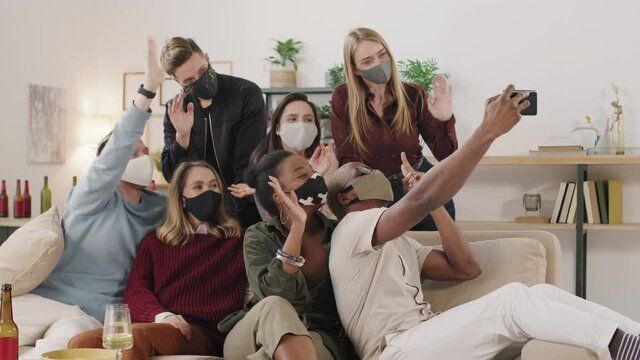 PAN slowmo of group of young men and women in face masks taking selfie on mobile phone at get-together in apartment during pandemic