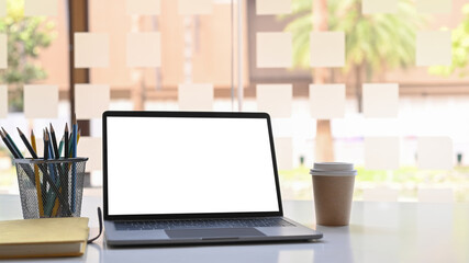 Open computer laptop with blank screen, notebook, coffee cup and pencil holder on white table near window.