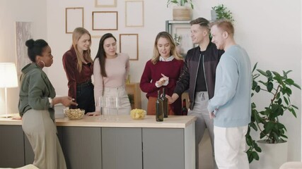 Medium shot of happy young man entering frame and putting bottles of wine on counter while his group of friends chatting at housewarming party or get-together
