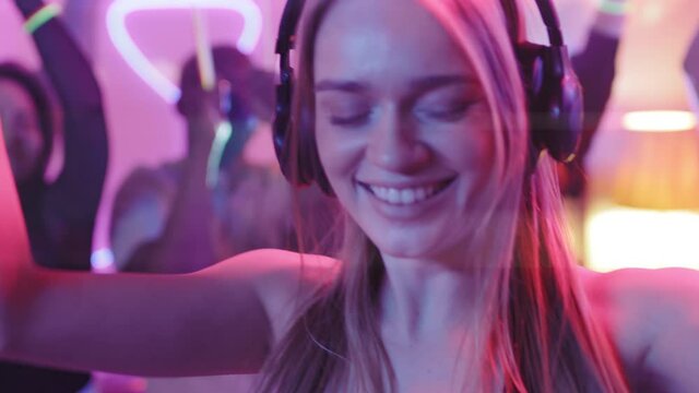 Close up shot of happy young woman in headphones DJing and dancing with group of young people at party