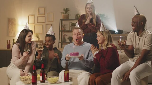PAN shot of diverse group of friends surprising their male friend with birthday cake. They are cheering and clapping while he is blowing candle, then hugging and congratulating him