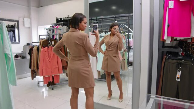 Fashionable african female in a clothing store chooses a raincoat, stands near the mirror and photographed on a smartphone, woman takes a selfie and shopping for clothes.