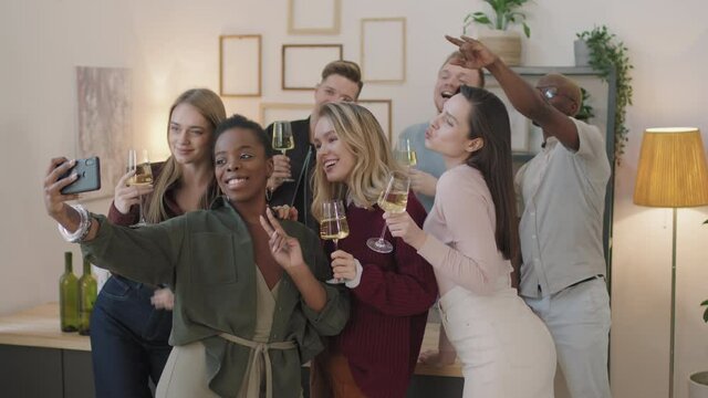 Medium shot of happy group of young friends holding glasses filled with champagne or white wine and posing for selfie at house party