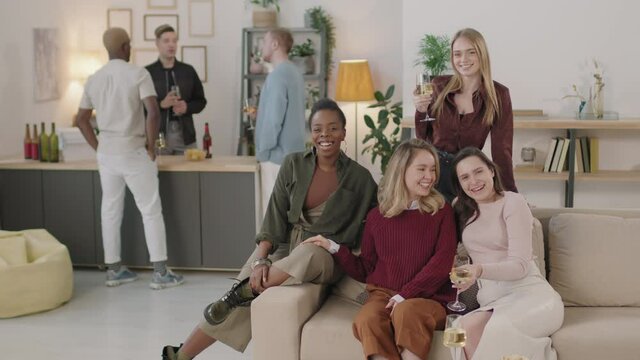 Slowmo shot of happy young women laughing and posing on couch in apartment at get-together. Three men chatting in background