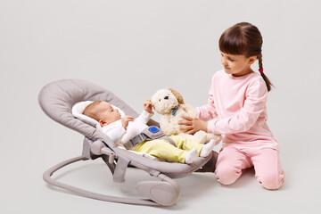 Happy sibling posing together isolated over white background, elder sister showing soft dog toy to little cute baby lying in rocking chair, happy childhood.
