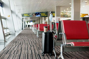 Next to the seats in an empty airport is a suitcase, on it is a glass of coffee, hanging a medical protective mask. Coronavirus, social distance, isolation