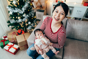 happy first-time mom looking up at camera while feeding her baby. view from above asian female seated on couch and holding her hungry child.