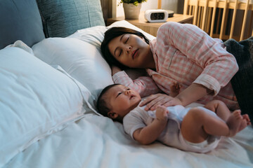 Obraz na płótnie Canvas tired asian new mom wearing pajamas is falling asleep on the bed while comforting her waking baby at night during bedtime in the bedroom.