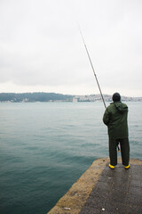 A lone mid age fisherman with yellow boots fishing on the Bosphorus shore in Istanbul.