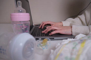 working mother-hand detail of a young mother working with her laptop on a white table with baby items in the home workplace.