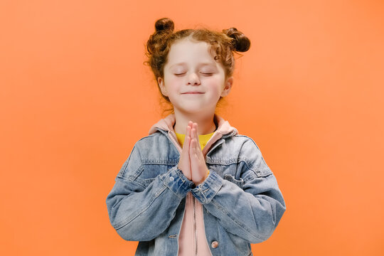Portrait of cute caucasian little girl kid praying with eyes closed, hands folded in prayer concept for faith, spirituality and religion, wears denim jacket, isolated on orange studio background