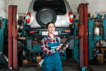 Portrait of young caucasian female mechanic in uniform posing with a tablet in her hands. In the background is an auto repair shop