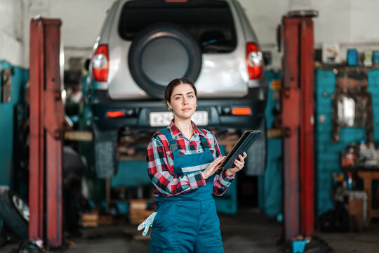 Portrait of young caucasian female mechanic in uniform posing with a tablet in her hands. In the background is an auto repair shop