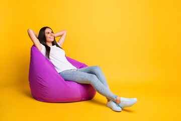 Photo of pretty charming young woman dressed white t-shirt sitting bean bag arms behind head isolated yellow color background
