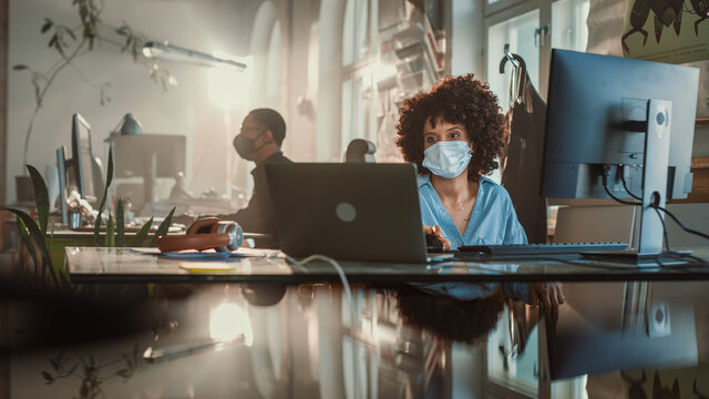 Creative Office: Black Woman Wearing Face Mask Sitting at Her Desk Working on Computer. Charmingly Authentic Professional with Curly Black Hair Creates Social Media Business Marketing Campaign