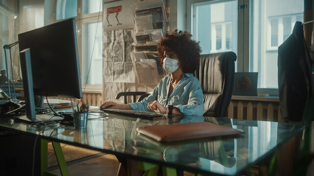 Young Black Woman Wearing Face Mask Working on Computer in Creative Modern Office. Authentic Content Creator, Game Software Developer, Marketing Specialist Creating Content. Medium Shot