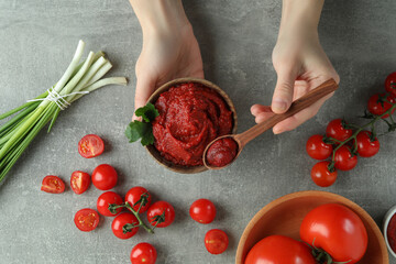 Female hands hold bowl and spoon with tomato paste on gray background with ingredients
