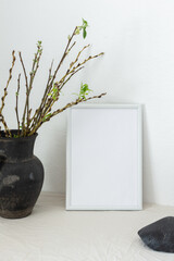 Home decor mock-up, blank picture frame near white painted concrete wall , black earthenware jug with pussy willow branches and blossoming green leaves