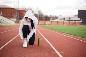 Obraz premium Young athletic girl tie shoelaces racetrack, prepares for a productive fitness workout on running track treadmill. Female sporty model dressed sportwear city stadium. Bottle shaker fresh water