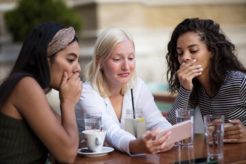 Three women sitting in café on street and talking. Using smart phone.