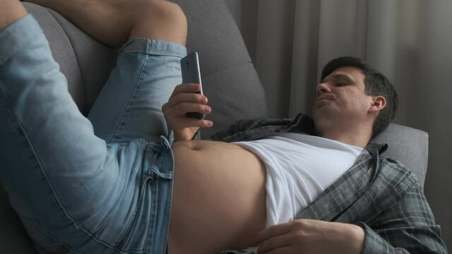Man is browsing smartphone lying on sofa at home with naked tummy. He is resting and scratching his belly. Looking on mobile phone screen in his free leisure time. Laughing and smiling.
