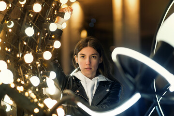 Millennial Hipster Girl Wearing Black Jacket Walking in City Street. Having Good Mood Over Glowing Festive Bokeh. Garlands Lights Urban Background.