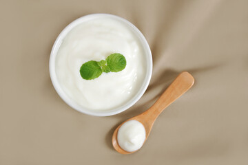 Top view of, Yogurt natural flavor in white ceramic bowl with peppermint leaf and wooden spoon placed on light brown cloth with copy space, Milk curd organic homemade for health care and healthy food.